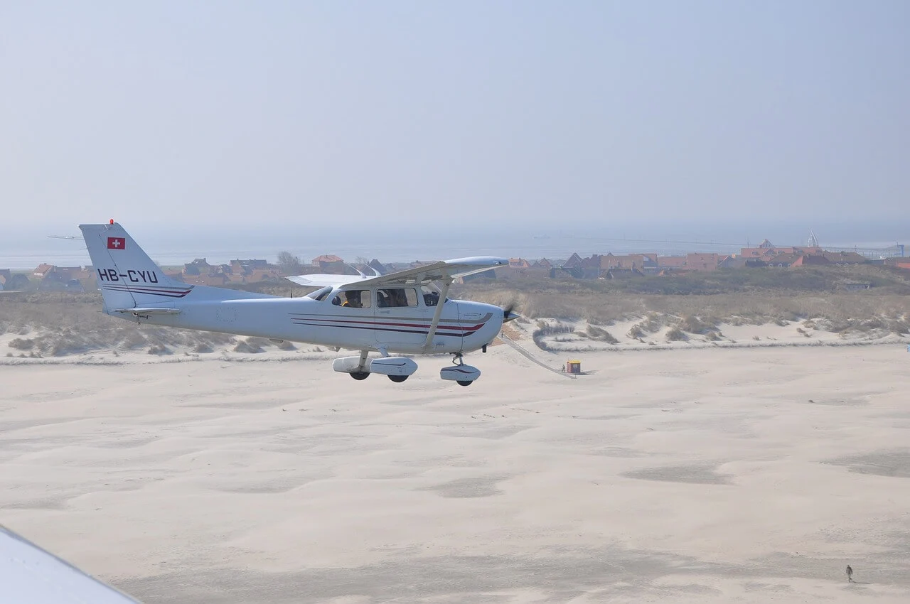 Ein Segelflugzeug mit schweizer Flagge am Flügel überfliegt einen Strandabschnitt. Im Hintergrund sind Häuser zu erkennen.