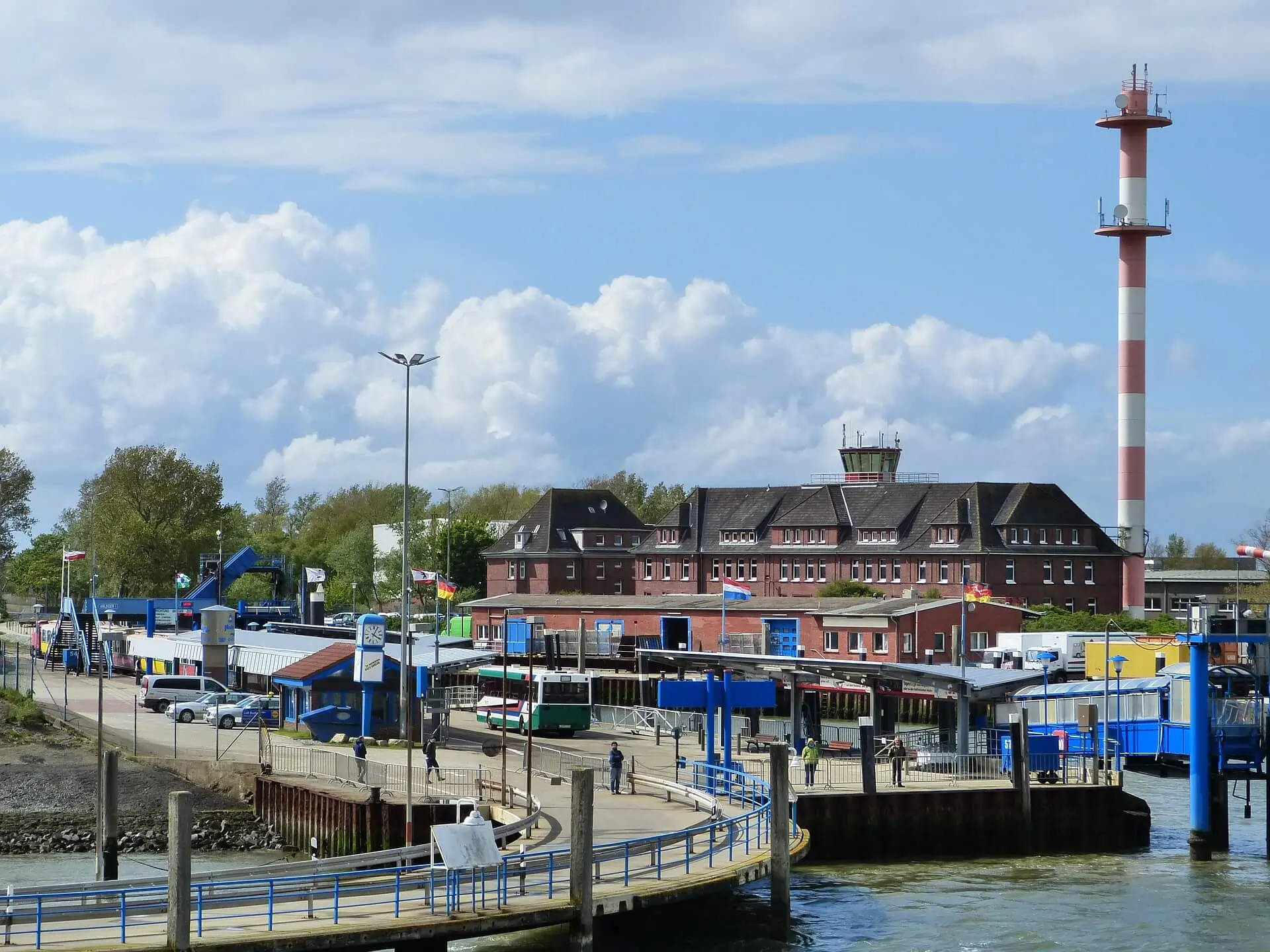 Der Borkumer Hafen mit dem Fähranleger auf der rechten Seite. Ein grün-weißer Bus wartet auf die Inselgäste. Im Hintergrund steht ein rotfarbenes Hafengebäude und ein rot-weißer Sendemast.
